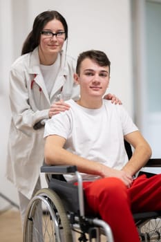 A smiling young man in a wheelchair is being pushed by a female healthcare worker in a white coat and stethoscope. They are in a hospital corridor, suggesting a medical or rehabilitation environment, evoking care and recovery.
