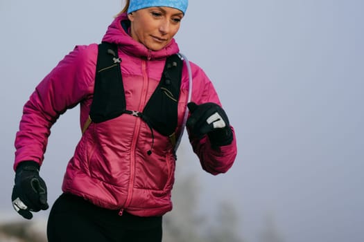 A woman athlete running along a rocky mountain trail in cold, misty weather, wearing a warm jacket, gloves, and a headband, showcasing endurance and determination.