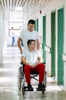 A smiling young man in a wheelchair is being pushed by a female healthcare worker in a white coat and stethoscope. They are in a hospital corridor, suggesting a medical or rehabilitation environment, evoking care and recovery.