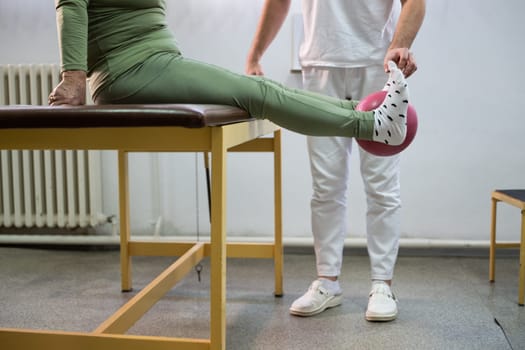 Close up of a patient performing a physical therapy exercise using a ball between the legs, assisted by a therapist.
