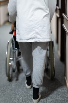 A smiling young man in a wheelchair is being pushed by a female healthcare worker in a white coat and stethoscope. They are in a hospital corridor, suggesting a medical or rehabilitation environment, evoking care and recovery.