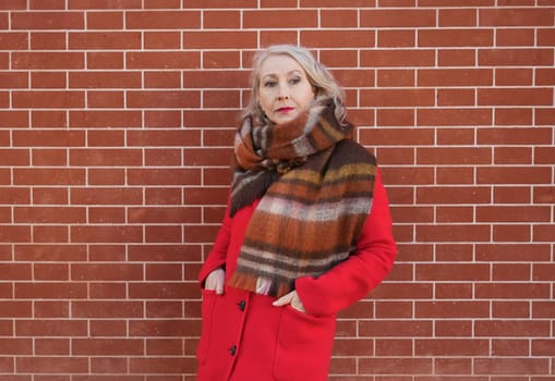 Gray-haired woman of sixty years old in a red coat and a terracotta scarf near a red -orange brick wall
