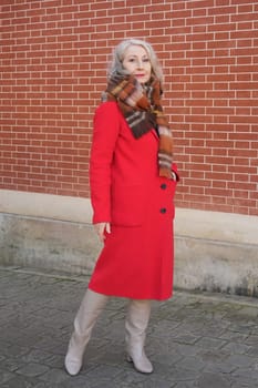 Gray-haired woman of sixty years old in a red coat and a terracotta scarf near a red -orange brick wall