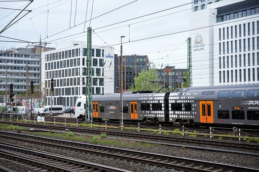 Dusseldorf, Germany 05.22.2025 Double decker commuter train moving past railway tracks with modern office buildings and overhead wires in background on a cloudy day