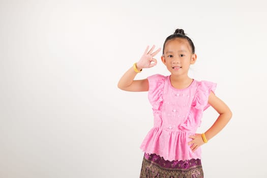 Portrait Thailand kid girl smiling traditional Thai dress costume showcases OK sign with smile, conveying agreement and positivity, studio isolated white background, kindergarten child success sign