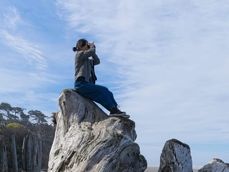 A middle aged woman looks out at the ocean with binoculars while sitting atop a large driftwood tree on a beach in Washington State.