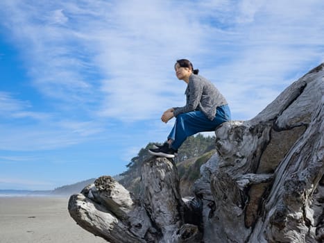 A middle aged woman sits on top of a large driftwood tree on the beach near Kalaloch area of Washington state.