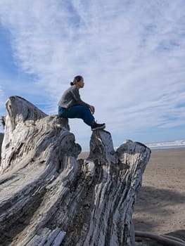 A middle aged woman sits on top of a large driftwood tree on the beach near Kalaloch area of Washington state.