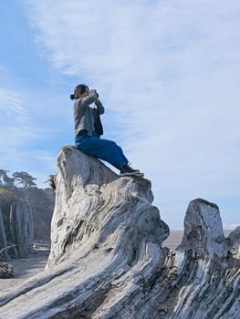 A middle aged woman looks out at the ocean with binoculars while sitting atop a large driftwood tree on a beach in Washington State.