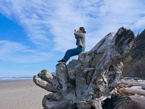 A middle aged woman looks out at the ocean with binoculars while sitting atop a large driftwood tree on a beach in Washington State.