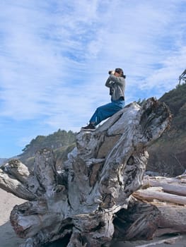 A middle aged woman looks out at the ocean with binoculars while sitting atop a large driftwood tree on a beach in Washington State.