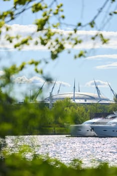 View of the mega yachts standing at the pier through the branches of trees on a clear sunny day, residential buildings and a stadium in the background, Russia, St.Petersburg. High quality photo