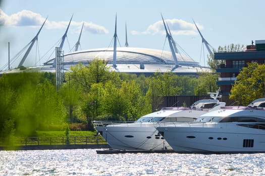 View of the mega yachts standing at the pier through the branches of trees on a clear sunny day, residential buildings and a stadium in the background, Russia, St.Petersburg. High quality photo