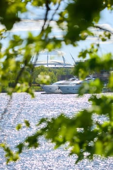 View of the mega yachts standing at the pier through the branches of trees on a clear sunny day, residential buildings and a stadium in the background, Russia, St.Petersburg. High quality photo