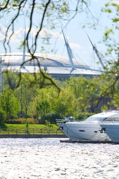 View of the mega yachts standing at the pier through the branches of trees on a clear sunny day, residential buildings and a stadium in the background, Russia, St.Petersburg. High quality photo