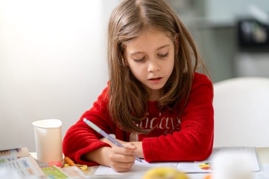 Cute girl in pajamas sitting at kitchen table, focused on doing homework. She is writing in notebook, surrounded by study materials, with calm and determined expression in cozy home environment