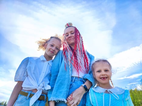 A mother and her two daughters enjoy a cheerful moment outdoors, surrounded by a bright blue sky and gentle clouds.