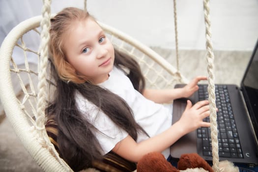 A young girl with long hair types on a laptop while sitting comfortably in a hanging chair, surrounded by soft toys.