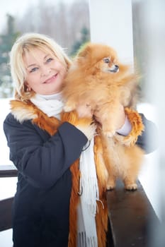 A middle-aged woman holds her small, fluffy red Pomeranian dog while walking in a snowy area during winter.