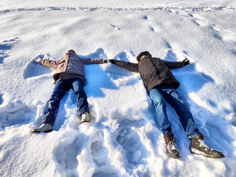 Two individuals lie on a snowy ground, creating snow angels in the bright winter sun. Their joyful expressions reflect the fun of winter activities amidst a serene snowy backdrop.
