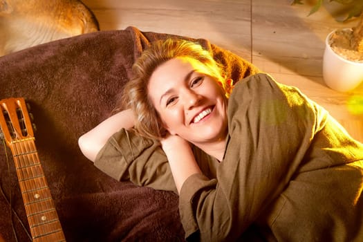 A woman lies back comfortably on a soft couch, smiling warmly with a guitar beside her in a naturally lit room.