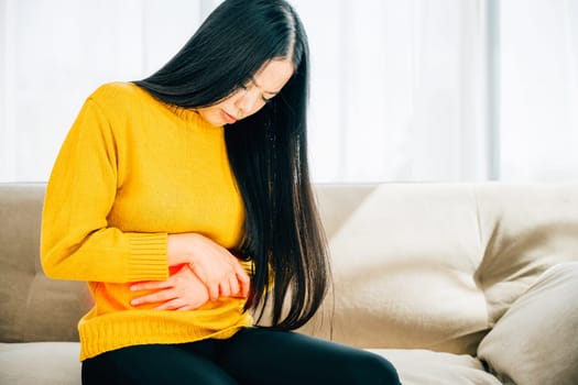 An Asian woman suffers from stomach ache on home couch in living room. Depicting discomfort symptoms and emphasizing the necessity of medical care for stomachache. Health care abdomen bloating concept