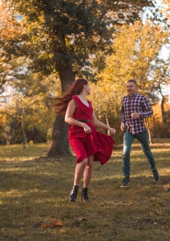 young couple in love actively relaxing in autumn park. they are running, having fun and laughing. girl in red dress against golden autumn background. concept of romance, falling in love.