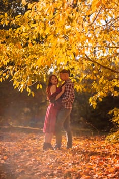 young couple in love hugging in autumn park, glare and sunlight around them. girl in red dress, against autumn trees. concept of falling in love, relaxation.