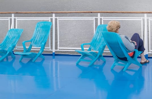 A lone middle-aged woman sits in a chaise lounge on the deck of a cruise ship, looking out to sea. Unrecognizable face.