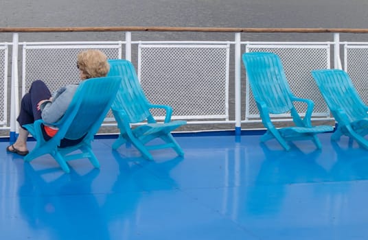 A lone middle-aged woman sits in a chaise lounge on the deck of a cruise ship, looking out to sea. Unrecognizable face.