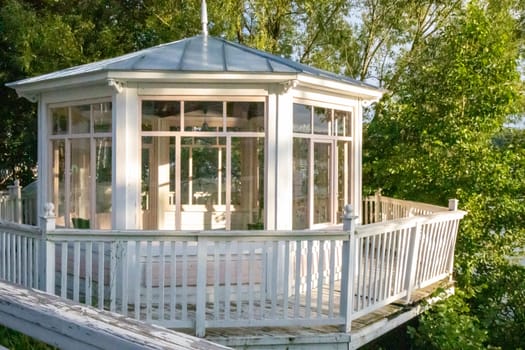 White wooden vintage gazebo with glass walls against a backdrop of green trees on a sunny summer day.
