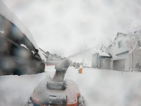 View from a snow-blanketed driveway captures a residential area mid-snowfall, with houses partially obscured by the cascading white flakes and a snow-covered car in the foreground.