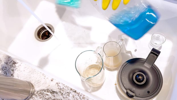Yellow-gloved hands are seen washing dishes, scrubbing diligently in a basin sink, surrounded by suds, cleaning utensils, and clear water glasses.