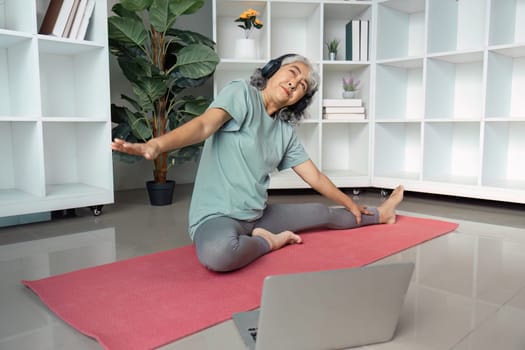 A senior woman stretches on a yoga mat, wearing headphones and engaging in an online yoga class, promoting wellness and active living.