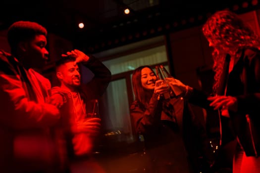 A group of friends share laughter and clink their beverage bottles in a vibrant atmosphere. The red lighting sets a cozy mood as they enjoy their time together.