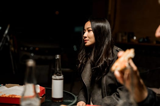 A young woman with long black hair wears a leather jacket while having pizza with friends at a nighttime gathering. The atmosphere is relaxed and casual.