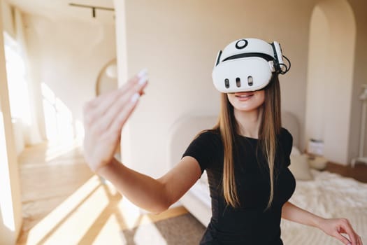 A young woman wearing a virtual reality headset interacts with a digital environment in a spacious, well-lit room. Sunlight streams in, highlighting her engagement with the technology.