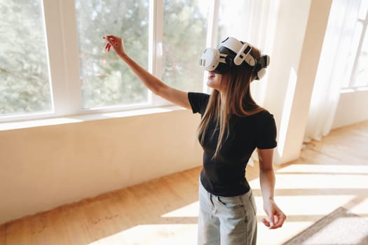 A young woman is fully immersed in virtual reality, wearing a headset and reaching out with her hand as sunlight filters through large windows. Wooden floors complement the relaxed atmosphere.