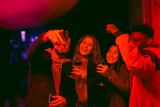 Friends enjoy a joyful celebration indoors during the night, sipping on drinks and taking selfies. They are surrounded by vibrant lighting, creating a lively atmosphere.