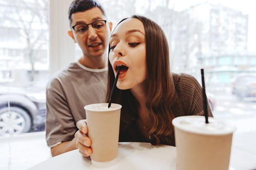 A young couple sits at a table in a warm cafe, smiling and sipping on their drinks. The rain falls outside, creating a cozy atmosphere. They are engaged in a fun and affectionate interaction.