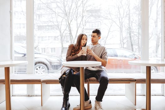 A couple shares playful moments while sipping coffee at a cafe. Frosted windows add a wintery backdrop to their cheerful interaction as they enjoy each other's company.