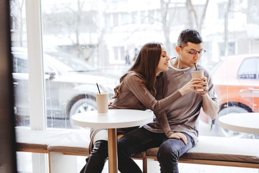 A young couple sits close together at a table in a cafe, sharing drinks and enjoying each other's company while snow falls outside.