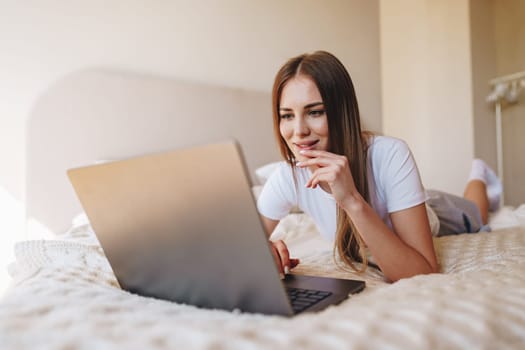 A woman with long hair is lying on her stomach on a bed, using a laptop. Natural light streams into the room, creating a cozy atmosphere as she engages with the screen.
