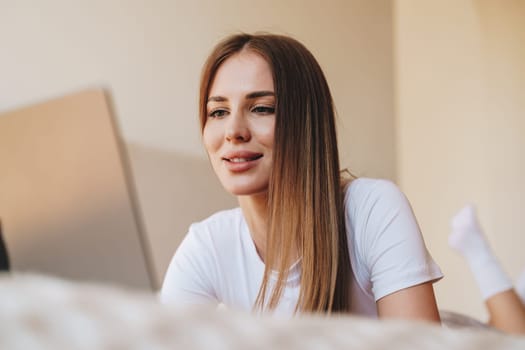 A young woman sits comfortably on a bed, focused on her laptop. Soft sunlight fills the room, creating a cozy atmosphere as she engages with her work.