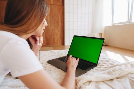 A woman is lying on her stomach on a bed, engaging with her laptop. The room is filled with natural light, creating a relaxed atmosphere perfect for work or leisure.