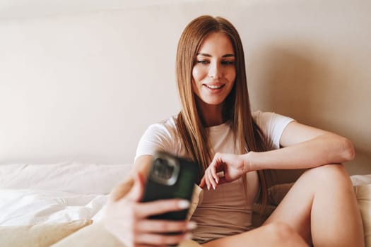 A young woman with long hair smiles as she engages in a video call, sitting comfortably on a bed with casual attire. The warm ambiance adds to her relaxed mood.