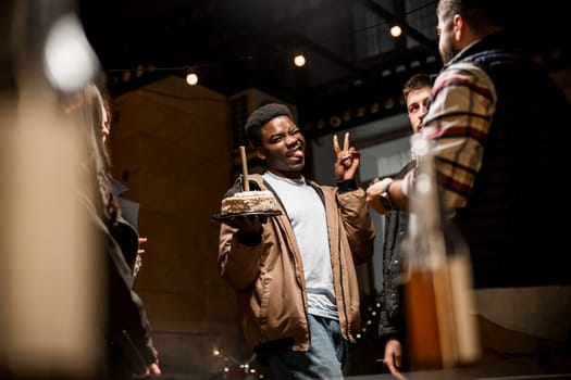 A group of friends gathers in a cozy setting, celebrating a birthday. One individual holds a cake, smiling and making a playful gesture. The atmosphere is festive and warm.