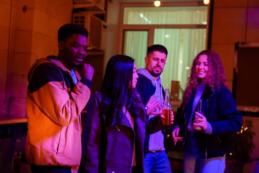 A group of four friends gather outdoors during a lively evening, engaging in conversation and enjoying drinks amidst bright, colorful lights, creating a festive atmosphere.