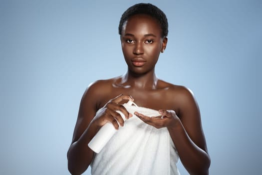 A young African woman with short hair, draped in a white towel, presses the dispenser of lotion into her hand. She's illuminated against a soft, gradient light blue background.