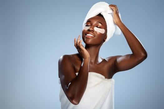 A woman is experiencing a spa treatment, smiling while applying facial patches. She is wrapped in a fluffy towel and has a towel on her head, enjoying self-care time.